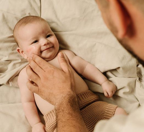 Bébé allongé sur un lot avec son père qui lui applique de la crème sur le visage - Mustela Benelux - 1