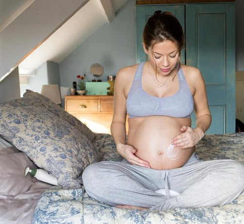 Femme enceinte assise sur un lit qui applique de la crème sur son ventre - Mustela Belgium - 1