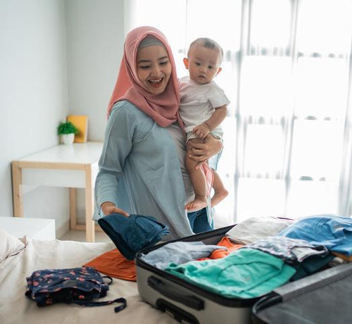 Maman qui tient son bébé dans ses bras et qui prépare une valise en mettant des vêtements à l'intérieur  - Mustela Belgium - 1