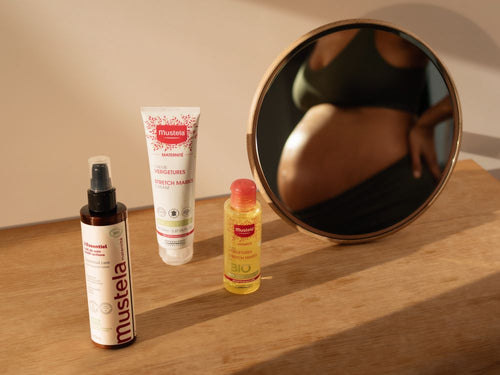 Produits de la gamme Maternité de chez Mustela posé sur une table, avec en fond le reflet dans un miroir d'une femme enceinte - Mustela Benelux - 6